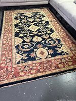 Full view of rectangular hand-knotted rug showing navy blue central field with floral vine motifs and rust-colored border, laid under sectional sofa on concrete floor.