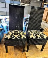 Front view of two black wicker patio chairs with white and black patterned cushions, showing woven wicker texture and wooden legs.
