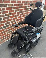 Front right three-quarter view of wheelchair against brick wall, shows seat, armrests, footrests and wheels.
