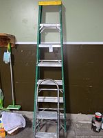 Full view of 7ft green fiberglass step ladder with aluminum steps and yellow top cap, placed in a garage with other items around.