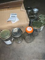 Multiple glass canning jars with metal lids inside and outside a cardboard box, shown from above and on floor.