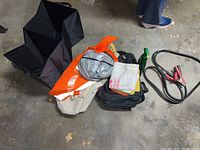 Collection of assorted car accessories laid on garage floor, showing jumper cables, safety vest, cleaning towels, organizer and spray bottle.