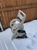 Light gray plastic hose reel with hand crank on the side, shown outdoors on snow with wooden fence background.