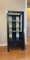 Front view of black media cabinet with frosted glass shelves, drawer at bottom, cable management holes visible on back panel.