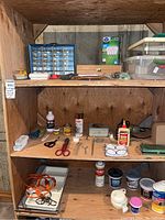 Three shelves with assorted repair and household items including adhesives, tapes, scissors, small parts, and containers.