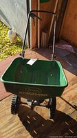Front view of green Scott's Standard broadcast spreader showing plastic hopper, two large black wheels, and metal handlebar on wooden floor with outdoor background.
