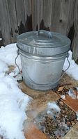 Photo of a small metal trash can outdoors on a wooden base surrounded by snow and dirt, showing the side view with lid and handle.