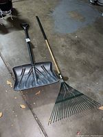 Photo of a black plastic snow shovel with metal handle and D-grip alongside a metal leaf rake with wooden handle and metal tines, both on a concrete floor with some dry leaves.