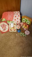 Photo showing a pink and red strawberry patterned sleeping bag alongside Strawberry Shortcake dolls, two framed pictures, and toy pieces.