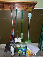 Photo showing multiple cleaning tools hanging and standing against a brown wall, including mops, duster, broom and dustpan, plus several cleaning bottles and cloths on the floor.