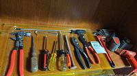 Photo showing an assortment of hand tools including screwdrivers, pliers, a red tape holder, and a few other tools on a wooden surface