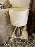 Single table lamp placed beside a couch showing the tear drop silver base and white drum shade on carpeted floor