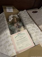 Porcelain Little Bo Peep doll partially wrapped in plastic, lying in opened Ashton Drake branded box with tissue paper. Certificate of authenticity visible in the box.