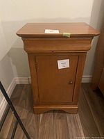 Solid wood bedside table, front view showing door and knob, wooden finish, placed on wooden floor.