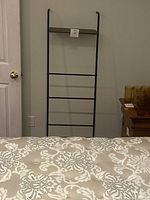 Photo of black metal blanket and quilt ladder leaning against wall near closed white door and small wooden table with amber glassware.