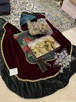 Lot items arranged on floor showing velvet burgundy tree skirt with black beaded fringe and gold trim, clear box with six gold bows, star tree topper, poinsettia floral Christmas stocking, and stocking hanger.