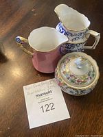 Three items shown on wooden surface including two small creamers and one sugar bowl with lid, all ceramic with hand painted designs and gold trim. MaxSold lot card visible.