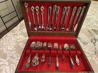 Wood box containing all flatware pieces arranged inside, showing multiple knife and fork designs with intricate handle detail.