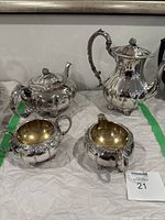 Photo showing front and side views of the two teapots, creamer, and sugar holder on a fabric background.