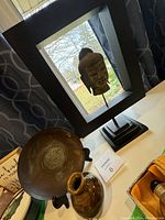 Photo showing a stoneware Buddha head statue mounted within a black frame on a wooden stand, with a vintage solid brass Chinese symbol plate and a Japanese vintage vase in front of it.