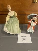 Photo showing both Royal Doulton figurines side by side on a flat surface, highlighting their size and appearance, with seller lot number label visible.