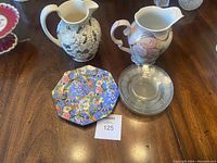 Lot overview showing two ceramic pitchers, one floral colorful octagonal plate, and one clear etched octagonal glass plate on wood table