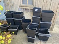 Black square and rectangular outdoor planters in three size groups, stacked and grouped on a concrete surface outside near a wooden fence.