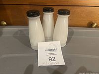 Three vintage milk glass shakers shown from front on a gray surface with wooden furniture background, black lids visible