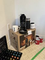 Nespresso Citiz Platinum & Milk coffee maker on top of its box along with milk frother, coffee mugs, and capsules case on floor beside it.