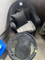 Black wicker patio armchair with striped cushion and two black throw pillows, next to matching black wicker circular table with glass top, placed indoors near a wall.