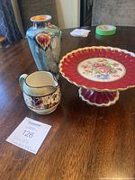 Cake plate with red scalloped edge, floral center, gold trim, displayed with vase and creamer pitcher on wooden table.