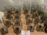 16 smoky tinted cocktail glasses with square bases, arranged in 4 rows of 4 on a wooden surface, showing the 'B' monogram on each glass.