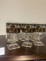 Front view of 5 Coca Cola stained glass style wine glasses showing square base, stem, and colorful branded rim.