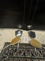 Two vintage silver hairbrush and hand mirror sets displayed upright on carpeted floor with patterned rug. Each set has one brush and one mirror showing the front sides.