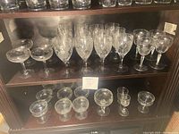 Photo of entire collection of etched glasses on two shelves showing large wine glasses, champagne coupe glasses, and smaller stemmed glasses.