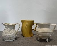 View of the full set of three creamers showing two off-white porcelain Belleek pieces and one yellow vintage glazed piece.