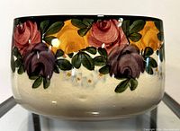 Front angle showing the floral design with purple, yellow, and reddish roses with green leaves along the top half of the vase, black rim and crackled white base.