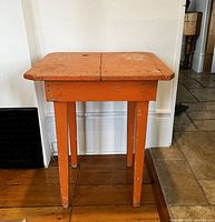 Front side view of antique pine table painted in orange showing full height and legs on wood floor.
