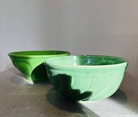 Two green ceramic serving bowls of different sizes and shades, shown together on a fabric surface with shadow and sunlight.