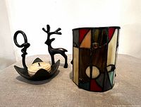 Photo showing three items in the lot: stained glass candle holder, steel reindeer figurine, handforged iron candleholder with beeswax candle on a table surface.