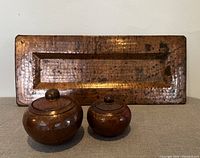 Front view of rectangular hammered copper plate with two round hammered copper lidded containers in front, showing texture and condition.
