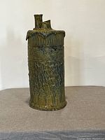 Tall handbuilt slab pottery vase with intricate textured pattern and glossy greenish-brown glaze, featuring three tubular openings on top.