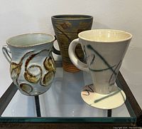 Three handmade pottery mugs including a tall abstract design, rustic textured mug, and small sculpted mug displayed on a glass surface.