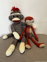 Front view of two sock monkeys sitting upright against wall. Classic one has grey and white knitted fabric with red pom-pom hat. Marileg version in orange and plaid patchwork body.