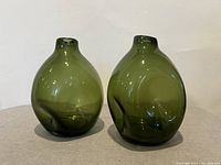 Pair of two olive green pinched glass vases shown side by side on light gray surface against white background under direct light, showing shape, color, and surface condition.