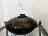 Close-up view of the raku oil burner pot showing curved shape, teal and brown glaze, and lid with wick.