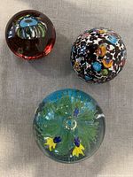 Three glass paperweights displayed together on grey fabric showing the aquarium style piece with fish, the large Millefiori piece, and smaller oval Millefiori piece