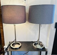 Two vintage table lamps placed side by side on a dark metal surface against a white wall, showing full lamp height and shade color.