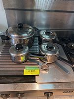 Four Lagostina stainless steel pots on a metal rack near a stovetop, all with black handles and knobs, showing the full set.