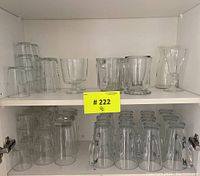 Assorted glass mugs, pitchers, and glasses arranged in a white kitchen cabinet with shelves.